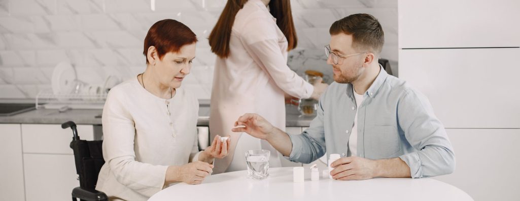 A caregiver assisting an elderly woman in a wheelchair with her medication at a kitchen table, while another person prep